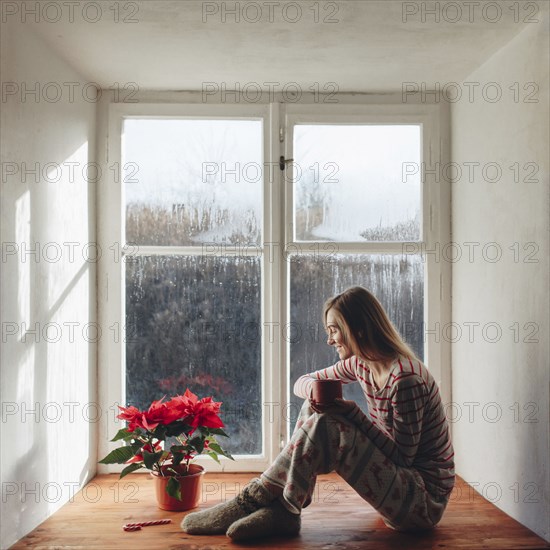 Caucasian woman sitting in window sill drinking coffee in pajamas