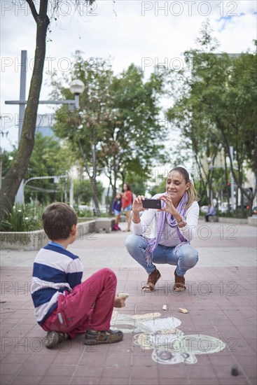 Hispanic mother photographing son on sidewalk