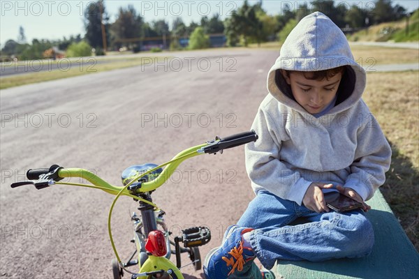 Hispanic boy sitting near bicycle texting on cell phone