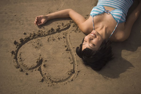 Asian woman laying next to heart in sand