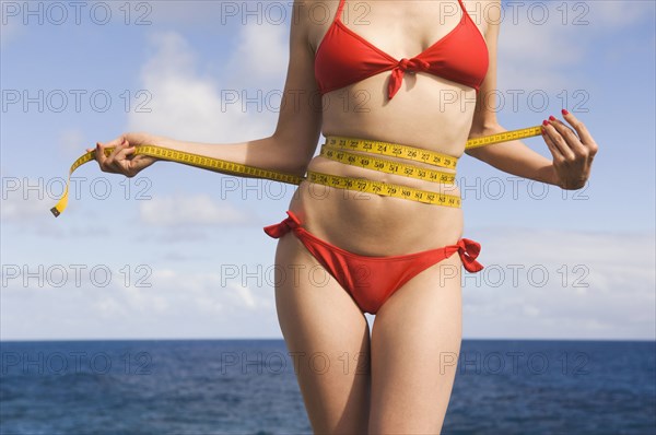 Mixed Race woman wearing bikini measuring waist near ocean