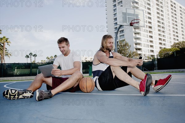 Caucasian men using cell phone and digital tablet on basketball court