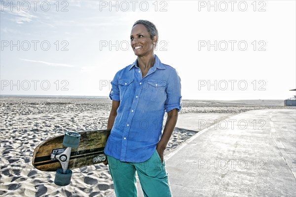 Older Black woman holding skateboard on beach