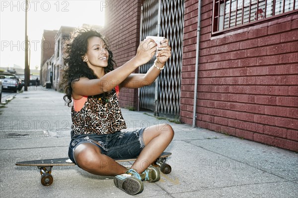 Hispanic woman taking cell phone picture on skateboard on city street