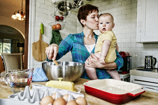 Caucasian mother kissing baby and cooking in kitchen