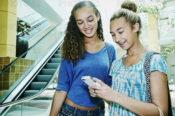 Mixed race teenage girls using cell phone near escalator at shopping mall