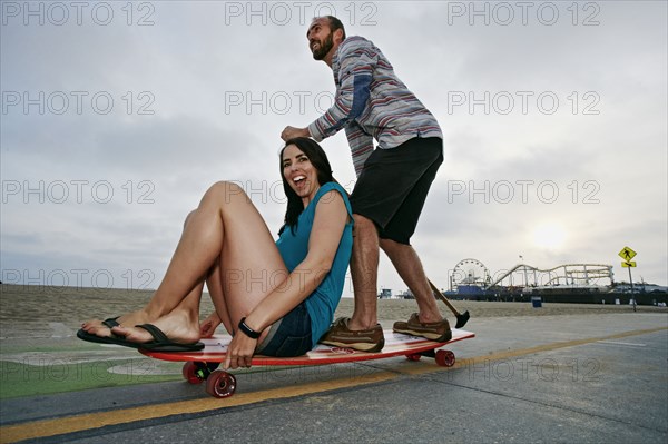 Caucasian couple skateboarding with paddle pole at beach