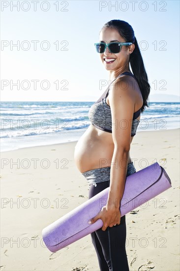 Pregnant Hispanic woman carrying yoga mat on beach