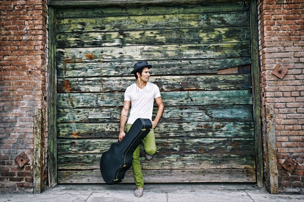 Mixed race musician holding guitar case near industrial building