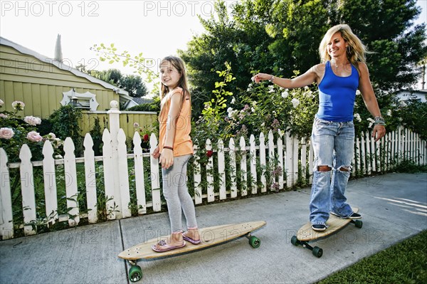 Caucasian mother and daughter riding skateboards on sidewalk