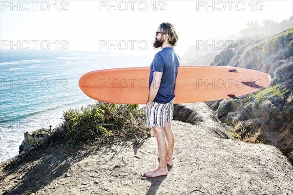 Caucasian man holding surfboard on coastal hilltop