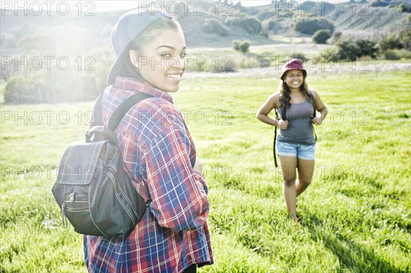 Smiling Mixed Race sisters backpacking in field near mountain