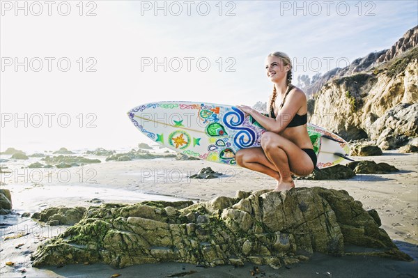 Caucasian woman crouching on rock carrying surfboard at beach