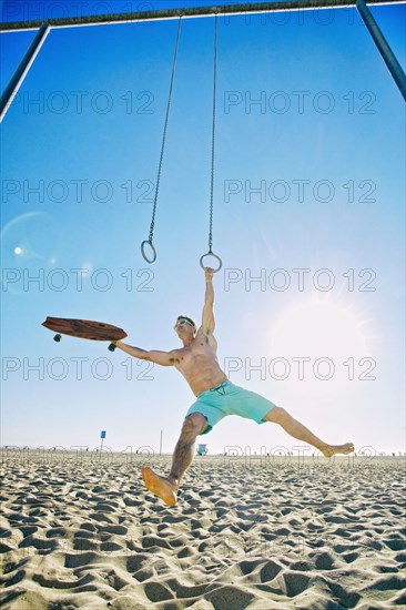 Caucasian man holding skateboard using gymnastic rings at beach