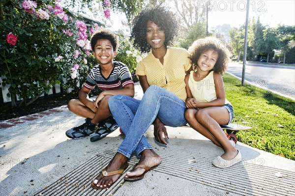 Smiling mother and children sitting on skateboard on sidewalk