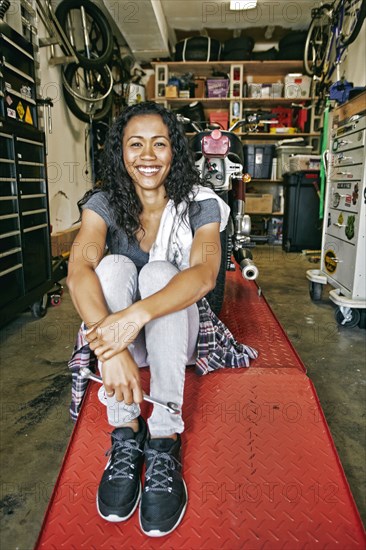 Smiling Mixed Race woman sitting on repair stand in garage