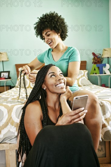 Woman braiding hair of friend holding cell phone in bedroom