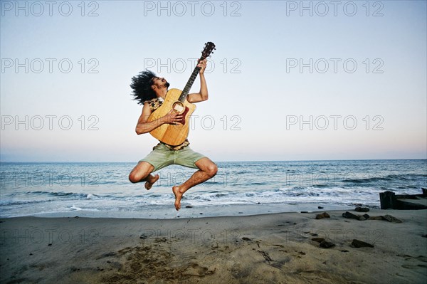 Mixed Race man playing guitar and jumping at beach