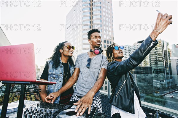 Men posing for cell phone selfie with DJ on urban rooftop