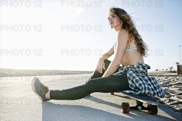 Caucasian woman sitting on skateboard on path at beach