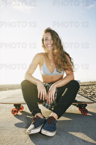 Caucasian woman sitting on skateboard on path at beach