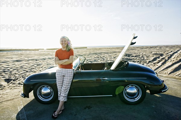 Older Caucasian woman leaning on convertible car with surfboard on beach