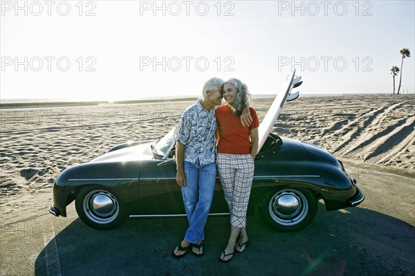 Older Caucasian couple leaning on convertible car with surfboard on beach