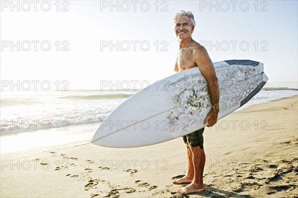 Older Caucasian man standing on beach carrying surfboard