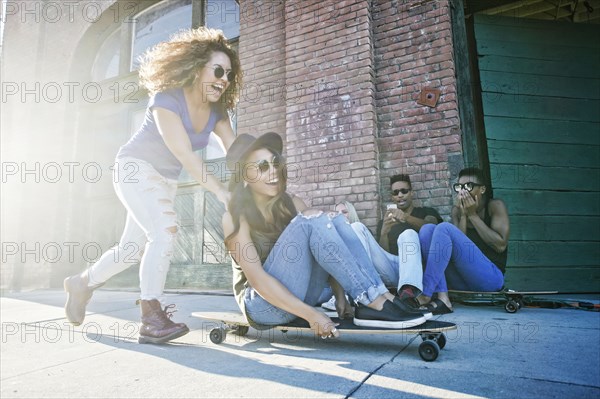 Woman pushing friend sitting on skateboard in city