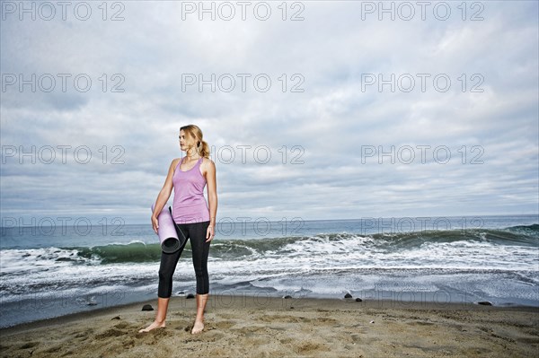 Caucasian woman standing on ocean beach holding exercise mat