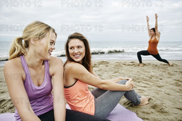 Caucasian woman performing yoga on beach near relaxing friends