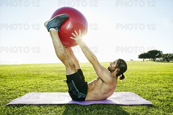 Caucasian man lifting fitness ball with legs in sunny field