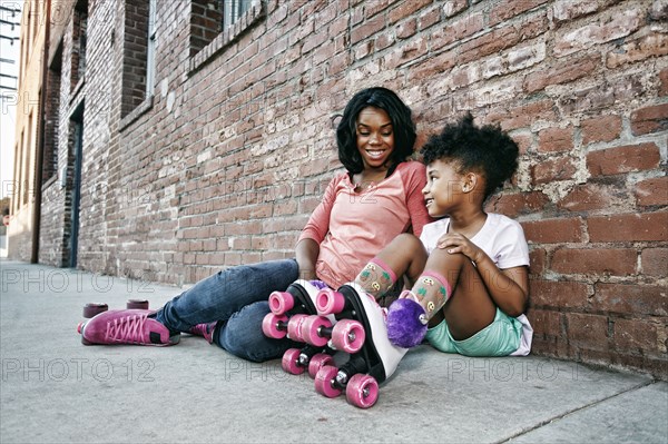 Black mother and daughter wearing roller skates sitting on sidewalk