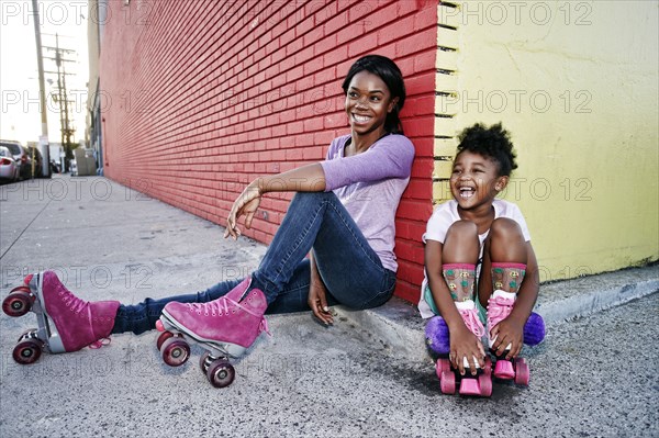 Black mother and daughter wearing roller skates sitting on sidewalk