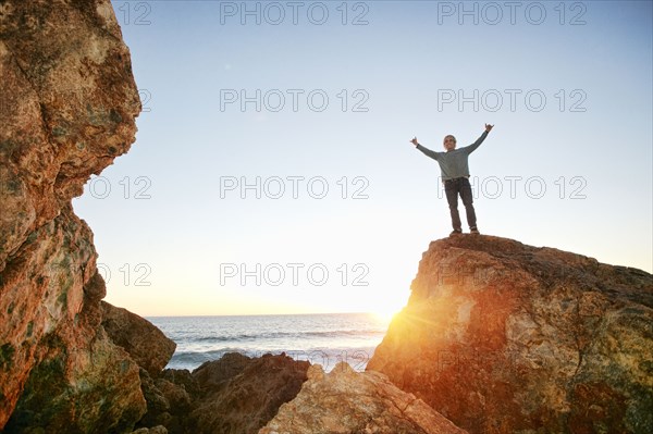 Caucasian man standing with arms raised on rock at ocean