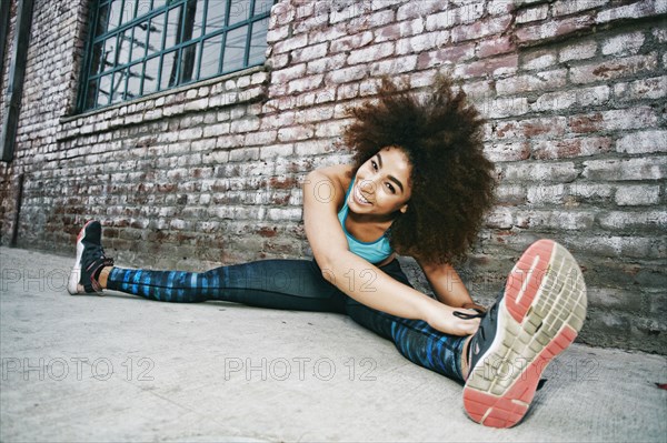 Hispanic woman sitting on sidewalk stretching legs near brick wall