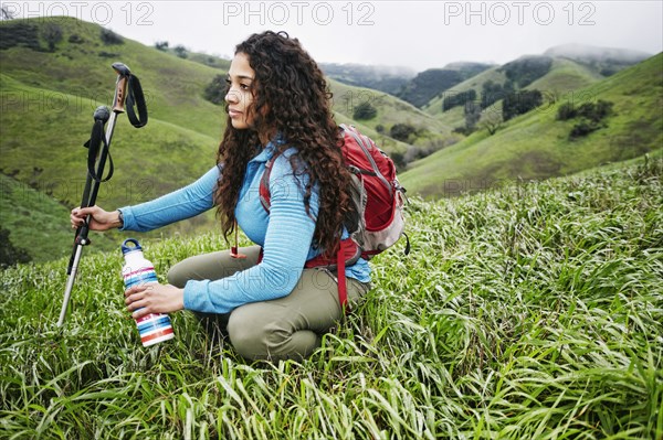 Mixed race woman crouching in grass holding walking sticks and bottle