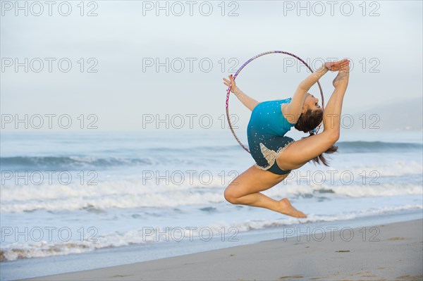 Caucasian gymnast jumping with hoop on beach