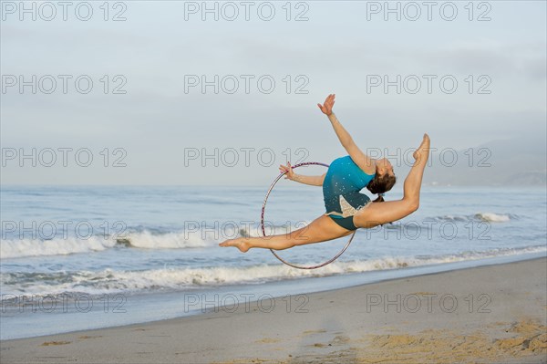 Caucasian gymnast jumping with hoop on beach
