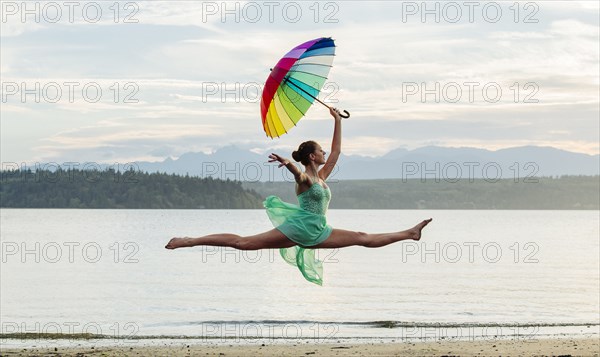 Caucasian ballerina jumping with multicolor umbrella on beach