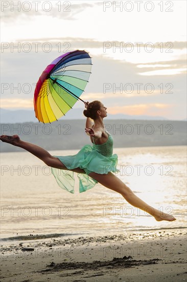 Caucasian ballerina jumping with multicolor umbrella on beach