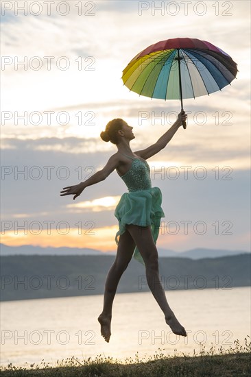 Caucasian ballerina jumping with multicolor umbrella on beach