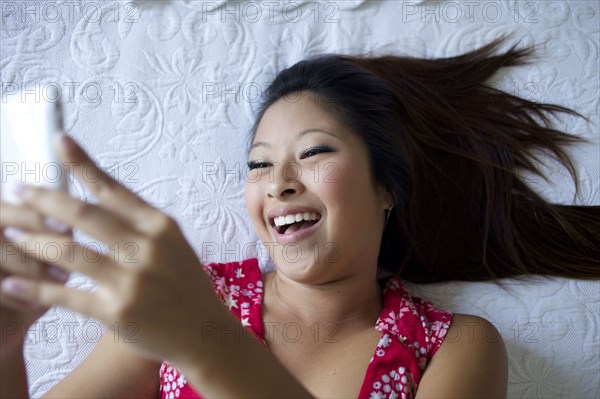 Smiling mixed race teenage girl laying on bed and text messaging on cell phone