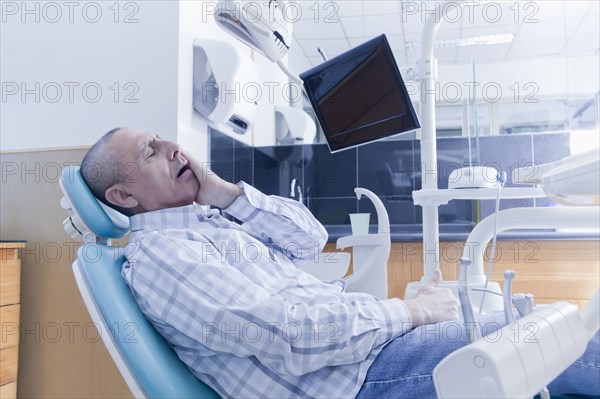 Older Hispanic man holding cheek in chair at dentist