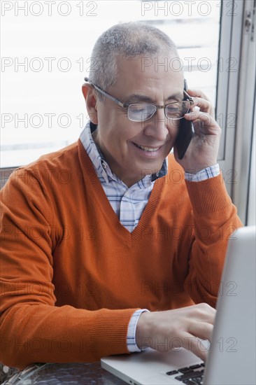 Smiling Hispanic man multi-tasking with cell phone and laptop