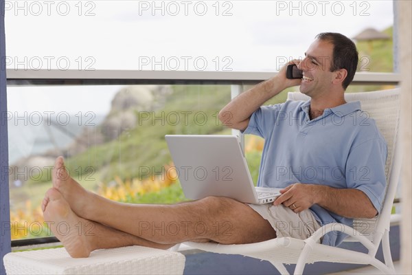 Man sitting on balcony with cell phone and laptop