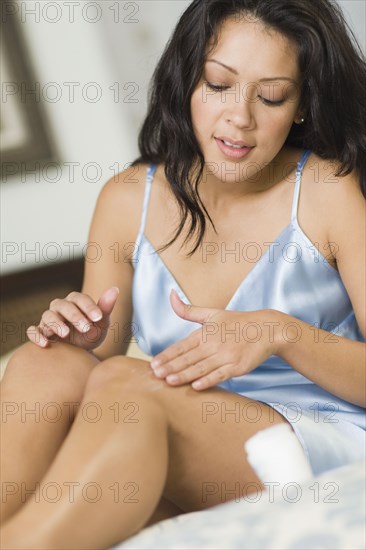 Hispanic woman rubbing lotion on legs in bedroom