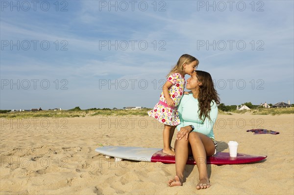 Girl kissing forehead of mother on surfboard on beach