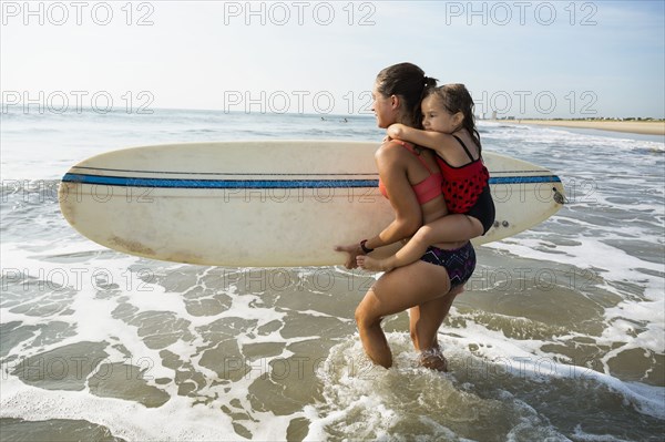 Mother carrying daughter and surfboard on beach