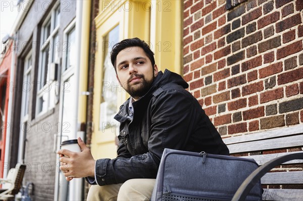 Hispanic man sitting on city bench drinking coffee